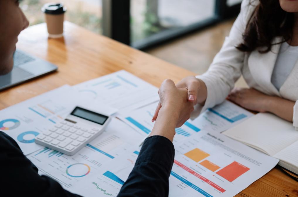“Two business professionals shaking hands across a desk covered with financial charts, documents, and a calculator, symbolizing a successful agreement or partnership for study visa process